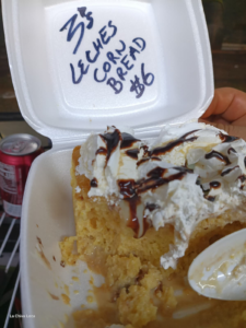 Slice of La Chiva Loca Colombian Tres Leches Corn Bread, soaked in milk, topped with rich whipped cream and dark chocolate sauce, served in a white container. Price $6.