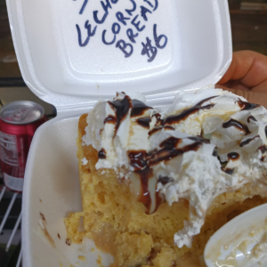 Slice of La Chiva Loca Colombian Tres Leches Corn Bread, soaked in milk, topped with rich whipped cream and dark chocolate sauce, served in a white container. Price $6.